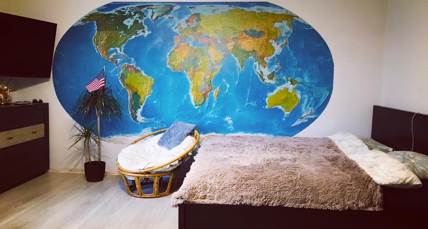 Pokój z dużym malowidłem ściennym przedstawiającym mapę świata w kształcie elipsy, obok łóżka i wiklinowego fotela, z małą flagą USA umieszczoną w doniczce z rośliną.