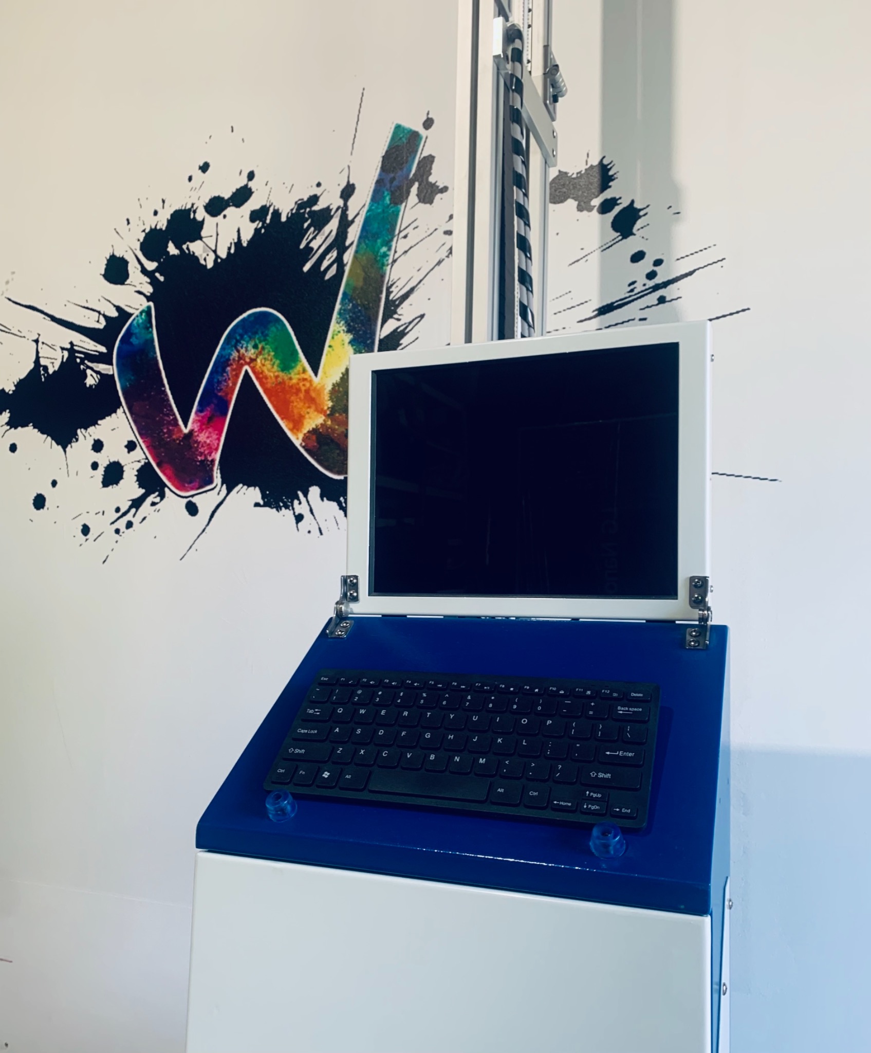 Urządzenie do druku na ścianach z widocznym ekranem, klawiaturą i częścią mechanizmu drukującego, obok ściana z kolorowym, abstrakcyjnym wzorem w kształcie litery W.