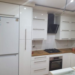 Art-dekor Artur Trela - Nowoczesna, biała zabudowa kuchenna z drewnianym blatem w Mielcu, obejmująca lodówkę Whirlpool, piekarnik i płytę gazową, z widocznymi elementami instalacji elektrycznej.