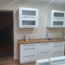 Nowoczesna kuchnia z białymi szafkami z szarymi szybami i drewnianym blatem, czajnik elektryczny i butelka wody na blacie, widok na wnętrze przez otwarte drzwi.