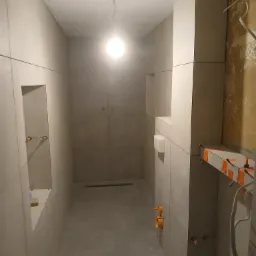 Wnętrze łazienki w trakcie prac wykończeniowych, widoczne szare płytki na ścianach i podłodze, przygotowane instalacje hydrauliczne, oraz fragment ściany przed położeniem płytek z widoczną starą...