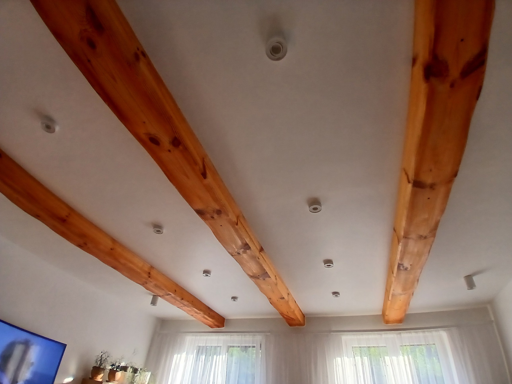 Jasne wnętrze z drewnianymi belkami stropowymi o nieregularnym kształcie i widocznymi sękami, oświetlone punktowymi lampami sufitowymi, z oknem w tle zasłoniętym białymi firankami i fragmentem...