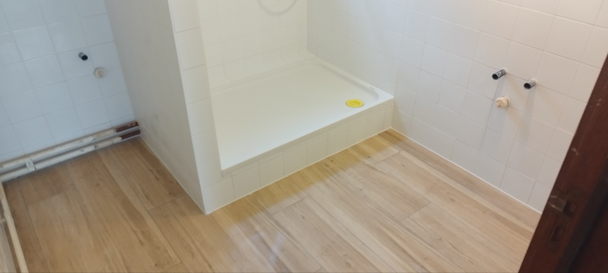 Biała kabina prysznicowa z brodzikiem, wyłożona białymi płytkami, z drewnopodobną podłogą i widocznymi rurami grzewczymi przy ścianie.