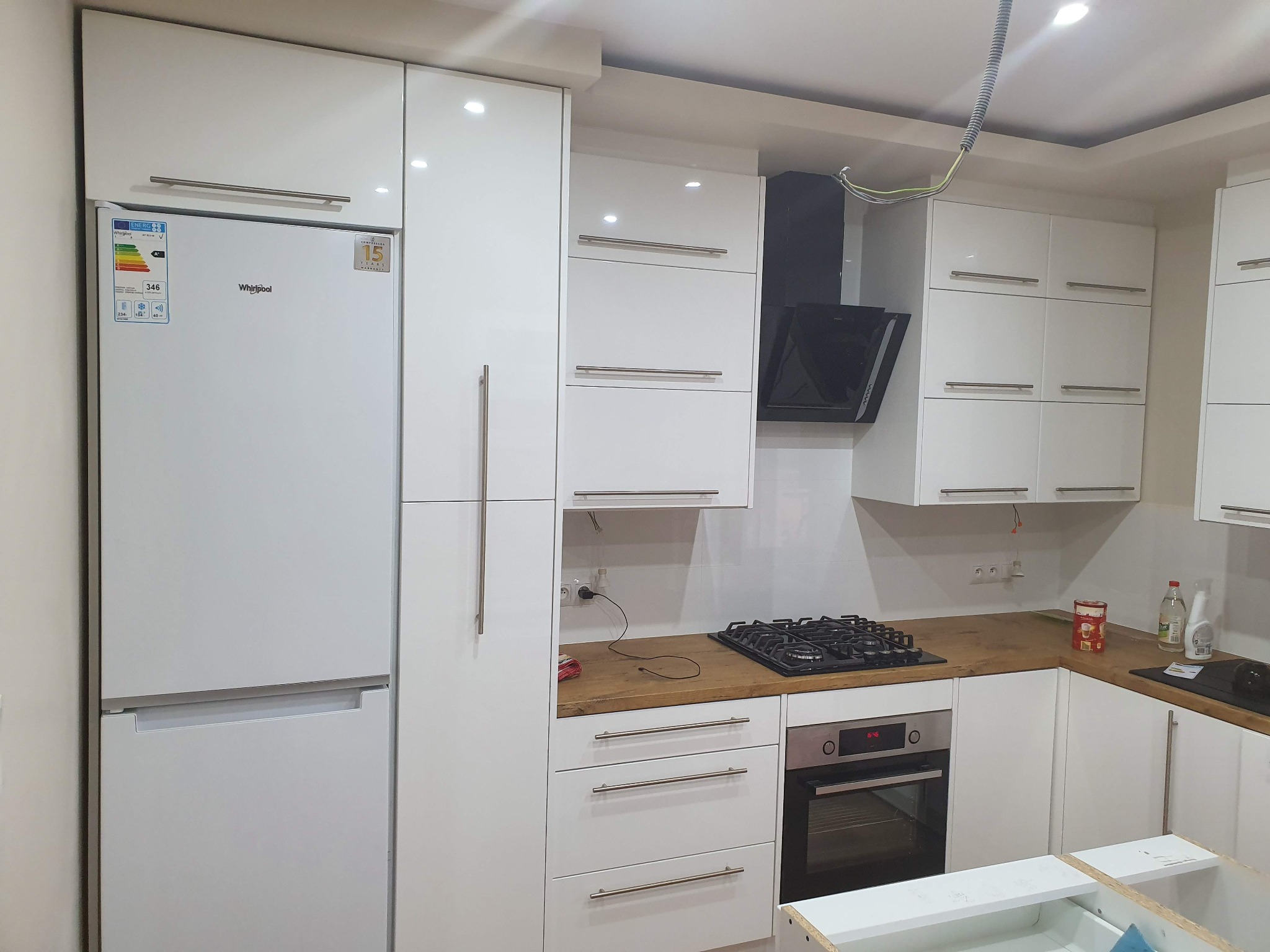 Nowoczesna, biała zabudowa kuchenna z drewnianym blatem w Mielcu, obejmująca lodówkę Whirlpool, piekarnik i płytę gazową, z widocznymi elementami instalacji elektrycznej.