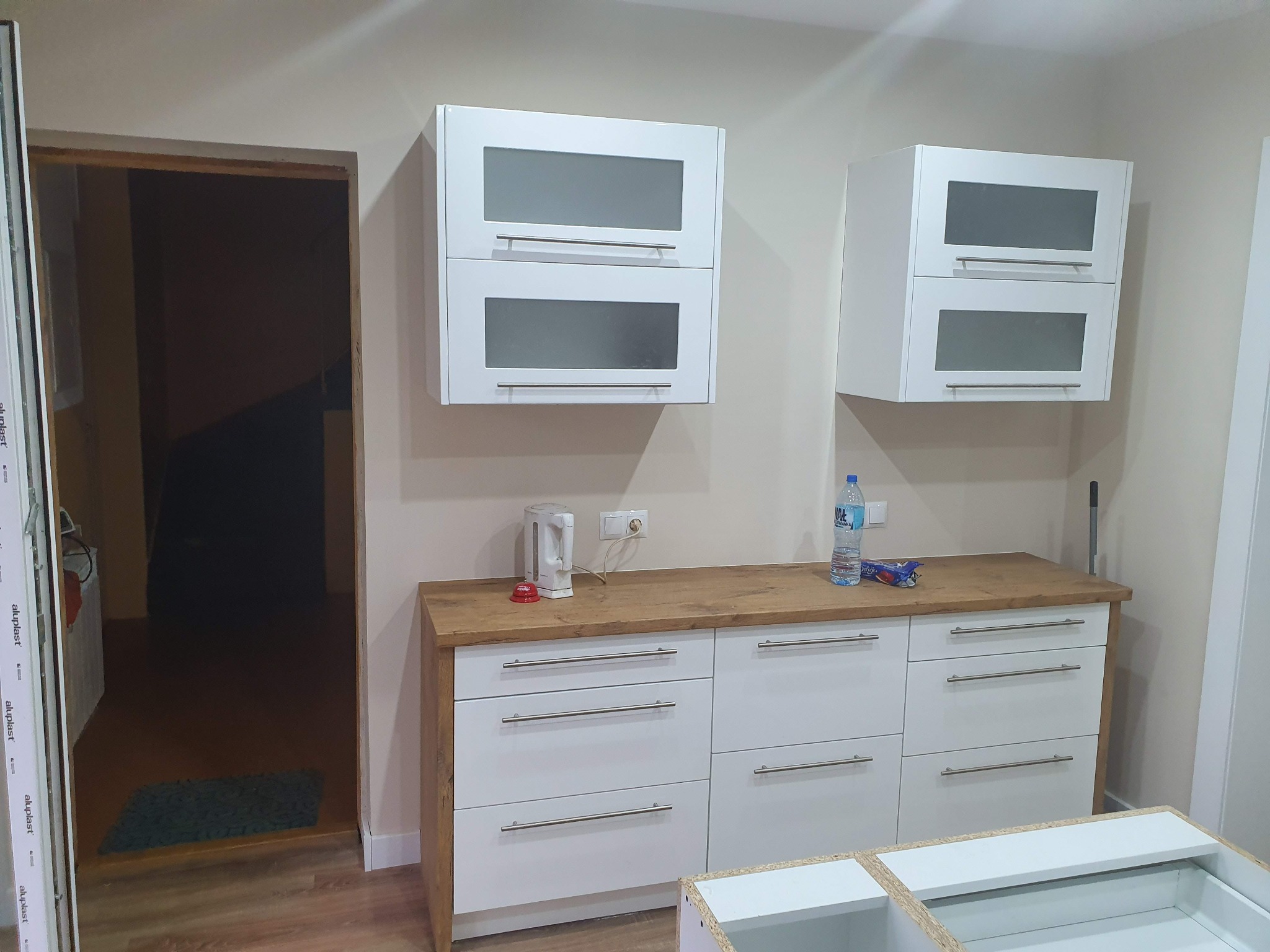 Nowoczesna kuchnia z białymi szafkami z szarymi szybami i drewnianym blatem, czajnik elektryczny i butelka wody na blacie, widok na wnętrze przez otwarte drzwi.