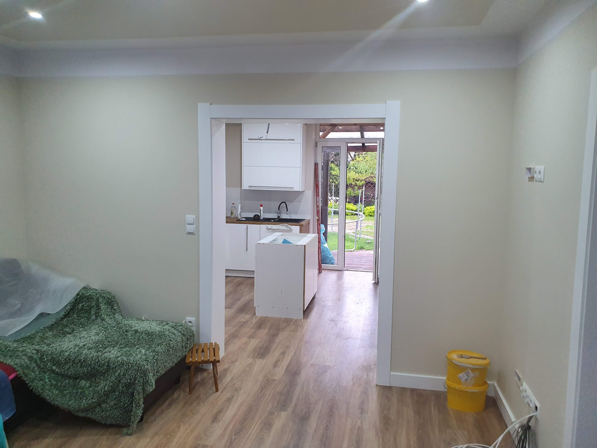 Widok z salonu na częściowo wykończoną kuchnię z białymi meblami, przez otwarte drzwi widać ogród, na podłodze panele, przy ścianie wiadro z farbą.