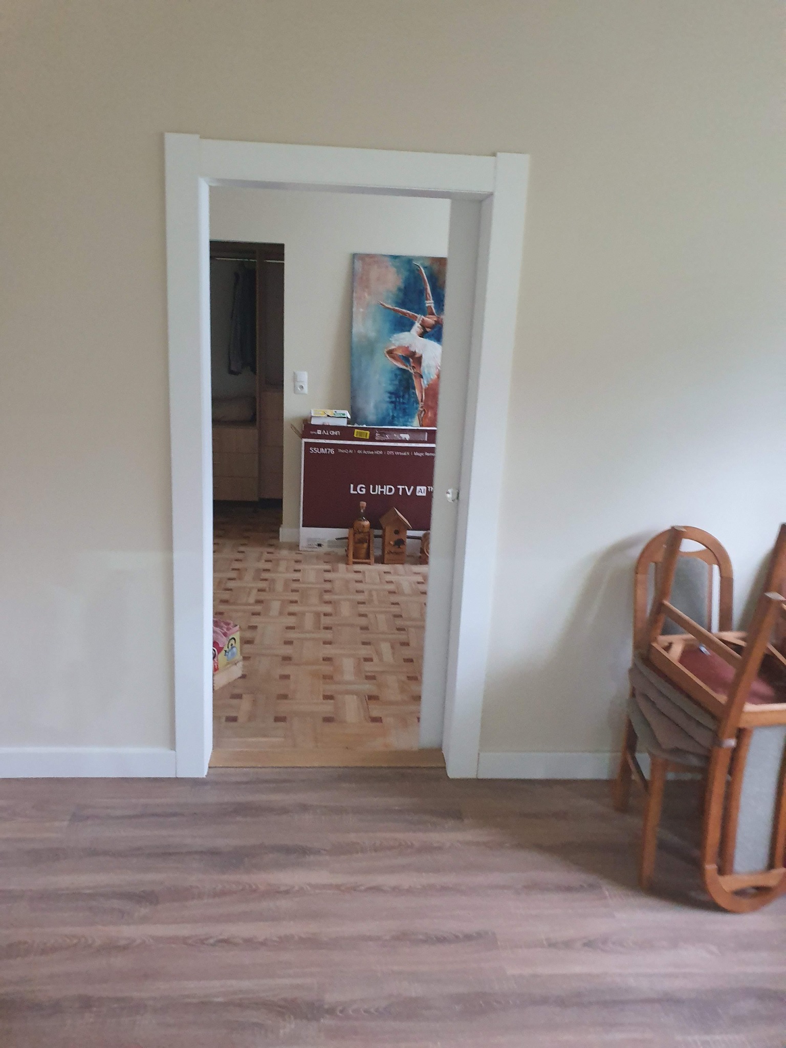 Widok na parkietową podłogę w pokoju przez nowo wykończone, białe futryny drzwiowe; w tle widoczny karton po telewizorze LG UHD TV i obraz baletnicy.