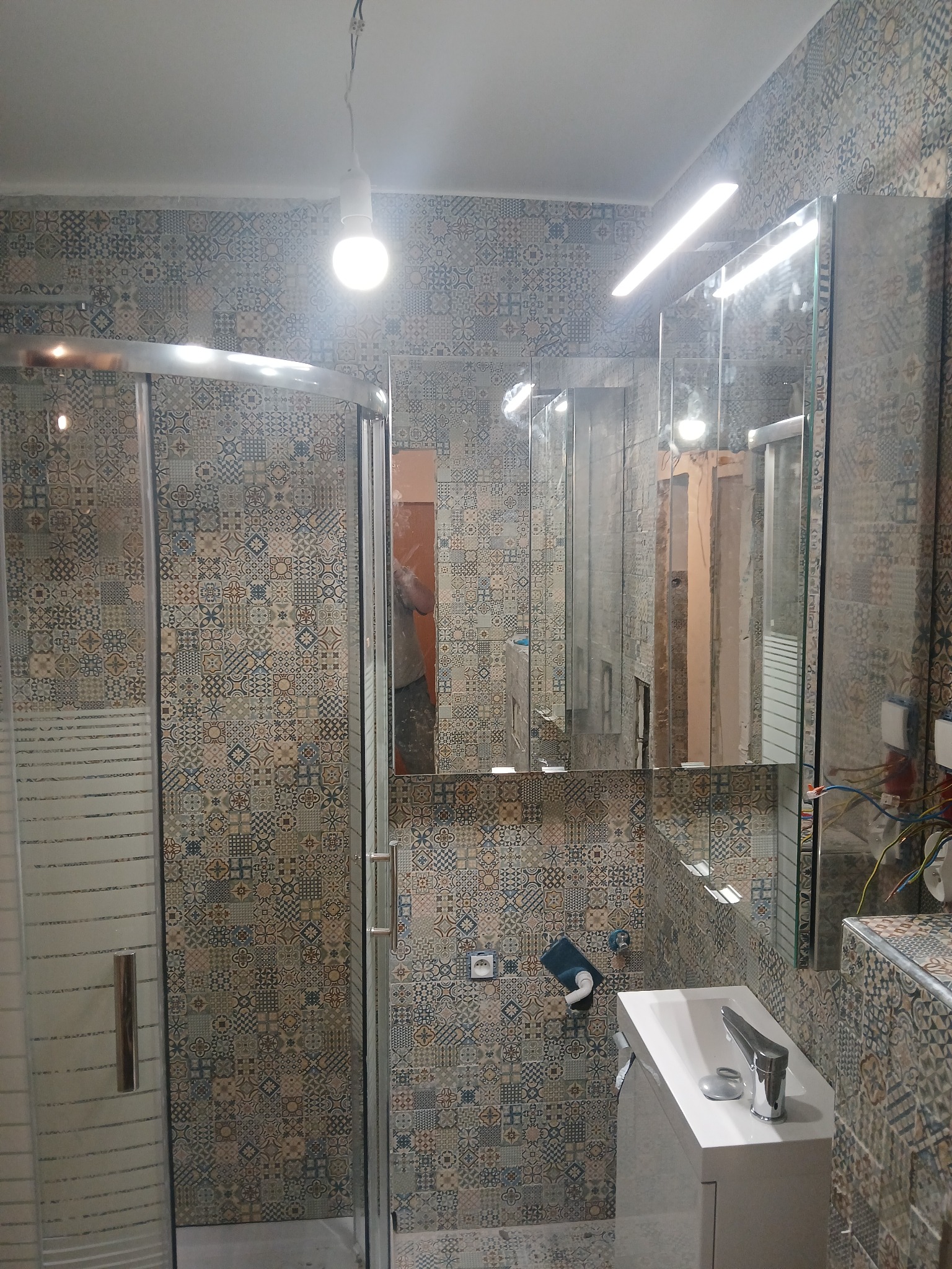 Ukończona łazienka z kabiną prysznicową, umywalką z szafką i lustrem, wykończona płytkami w patchworkowy wzór; widoczne niedokończone podłączenia elektryczne.