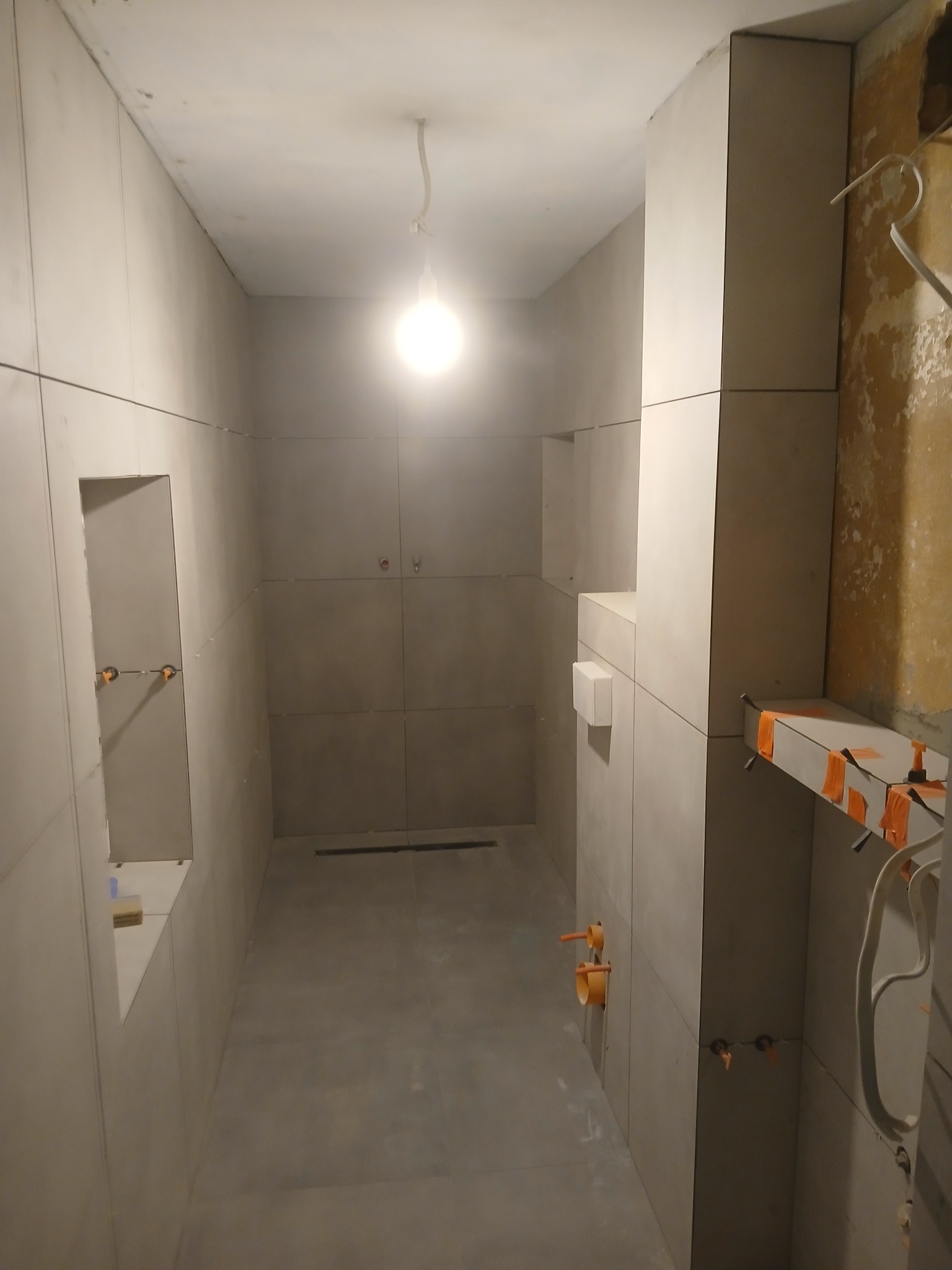 Wnętrze łazienki w trakcie prac wykończeniowych, widoczne szare płytki na ścianach i podłodze, przygotowane instalacje hydrauliczne, oraz fragment ściany przed położeniem płytek z widoczną starą...