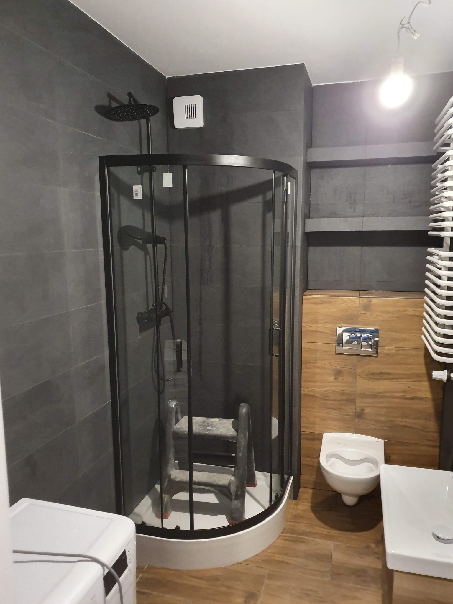Nowoczesna łazienka z kabiną prysznicową typu walk-in z czarnymi profilami, szare płytki na ścianach, drewnopodobne panele na ścianie z toaletą podwieszaną, drabinka grzejnikowa i pralka w rogu...
