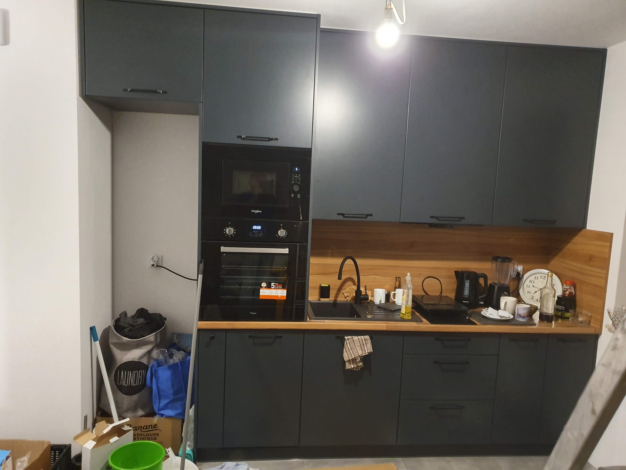 Szare szafki kuchenne z drewnianym blatem i oświetleniem punktowym, widoczny piekarnik Whirlpool z naklejką '5 lat gwarancji', w tle torba na pranie i resztki materiałów budowlanych.