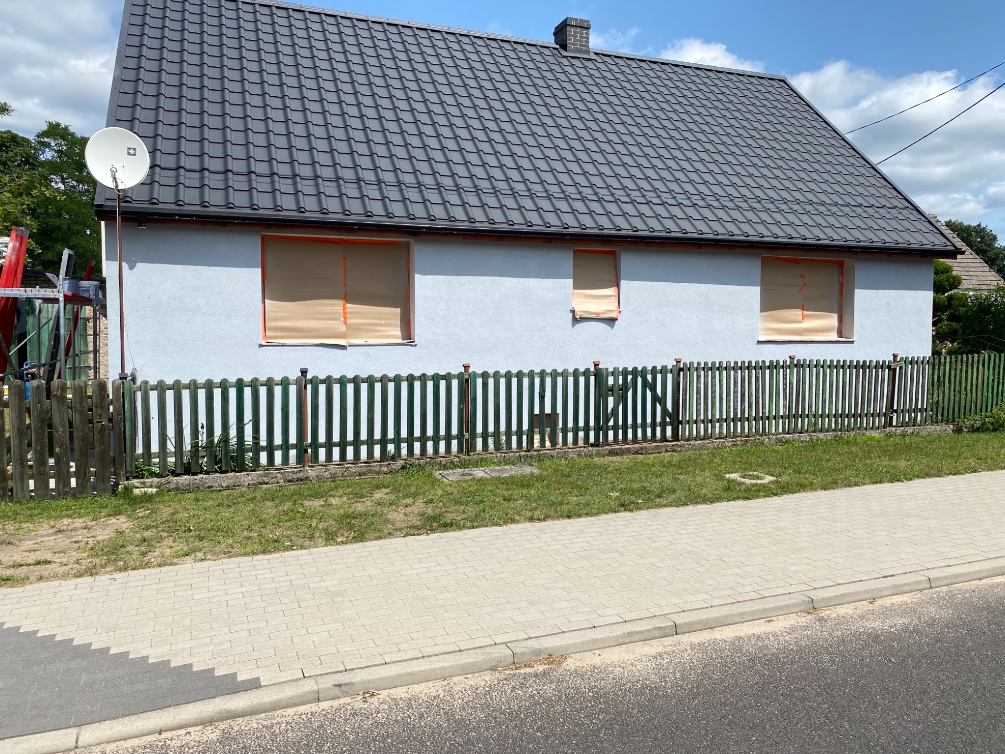 Dom jednorodzinny z szarą elewacją w trakcie przygotowań do prac wykończeniowych; okna zabezpieczone brązową folią malarską, widoczny fragment chodnika i zielonego ogrodzenia.