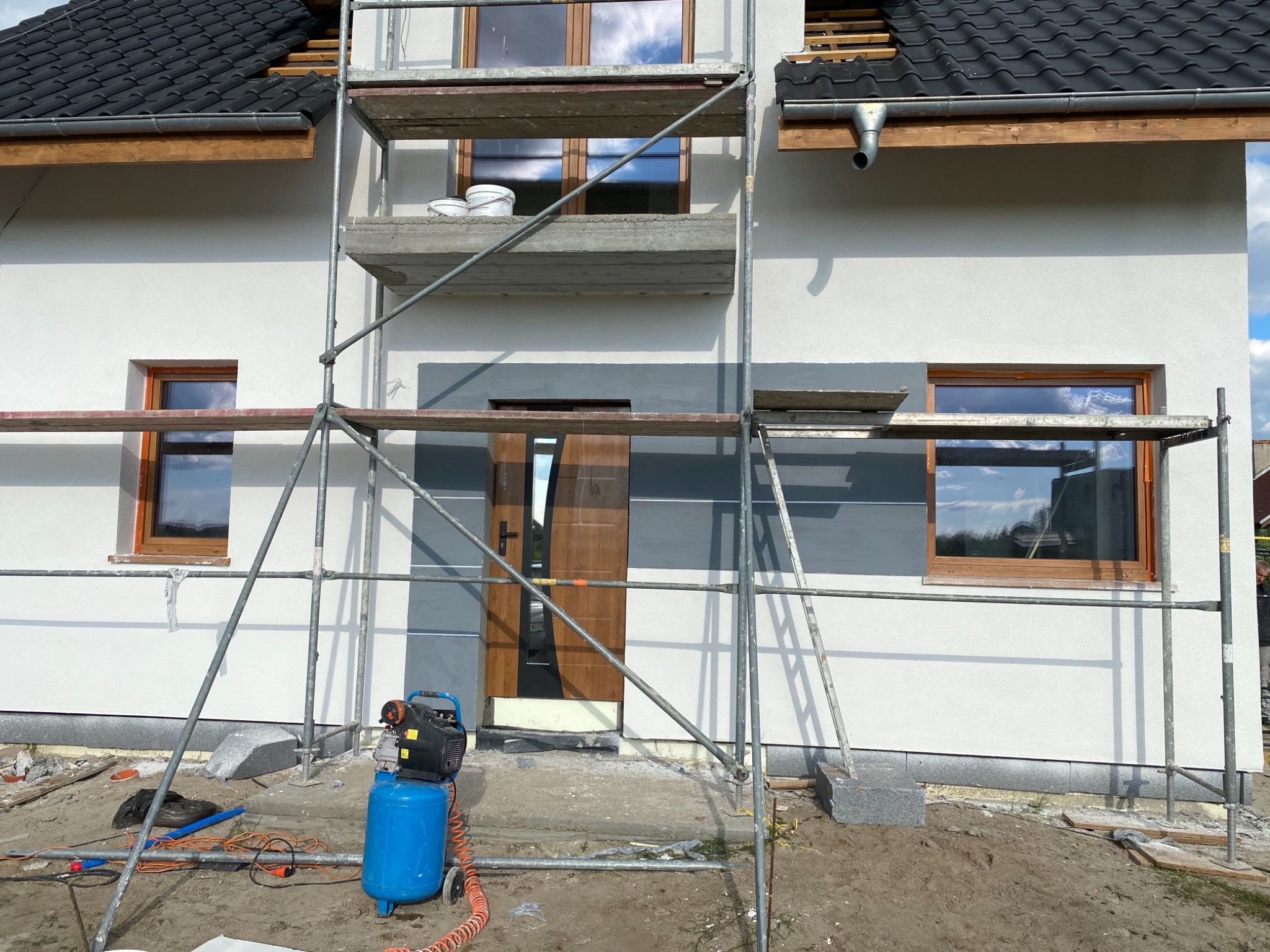Świeżo otynkowana elewacja domu jednorodzinnego, widoczne rusztowanie, kompresor pneumatyczny i częściowo zamaskowane okna przed malowaniem na szaro.