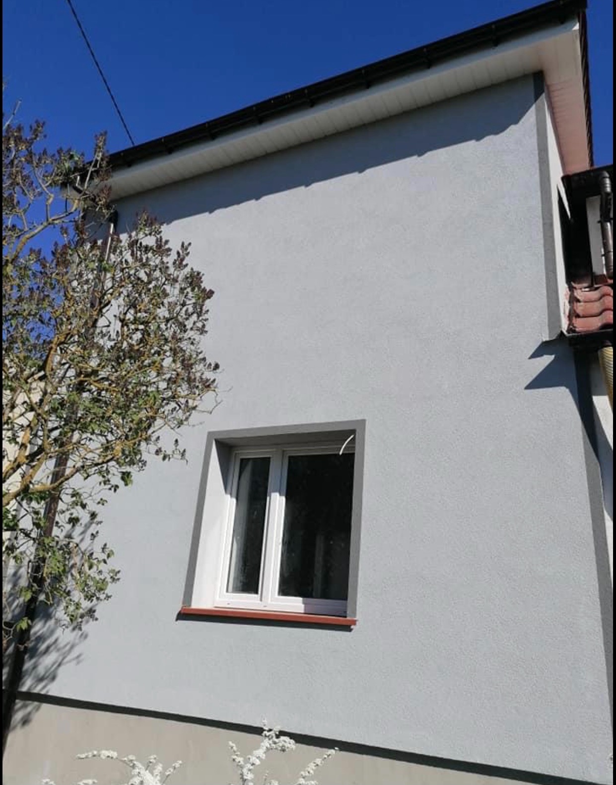 Jasnoszara elewacja budynku z oknem, widoczny fragment dachu i drzewo z boku, błękitne niebo w tle.