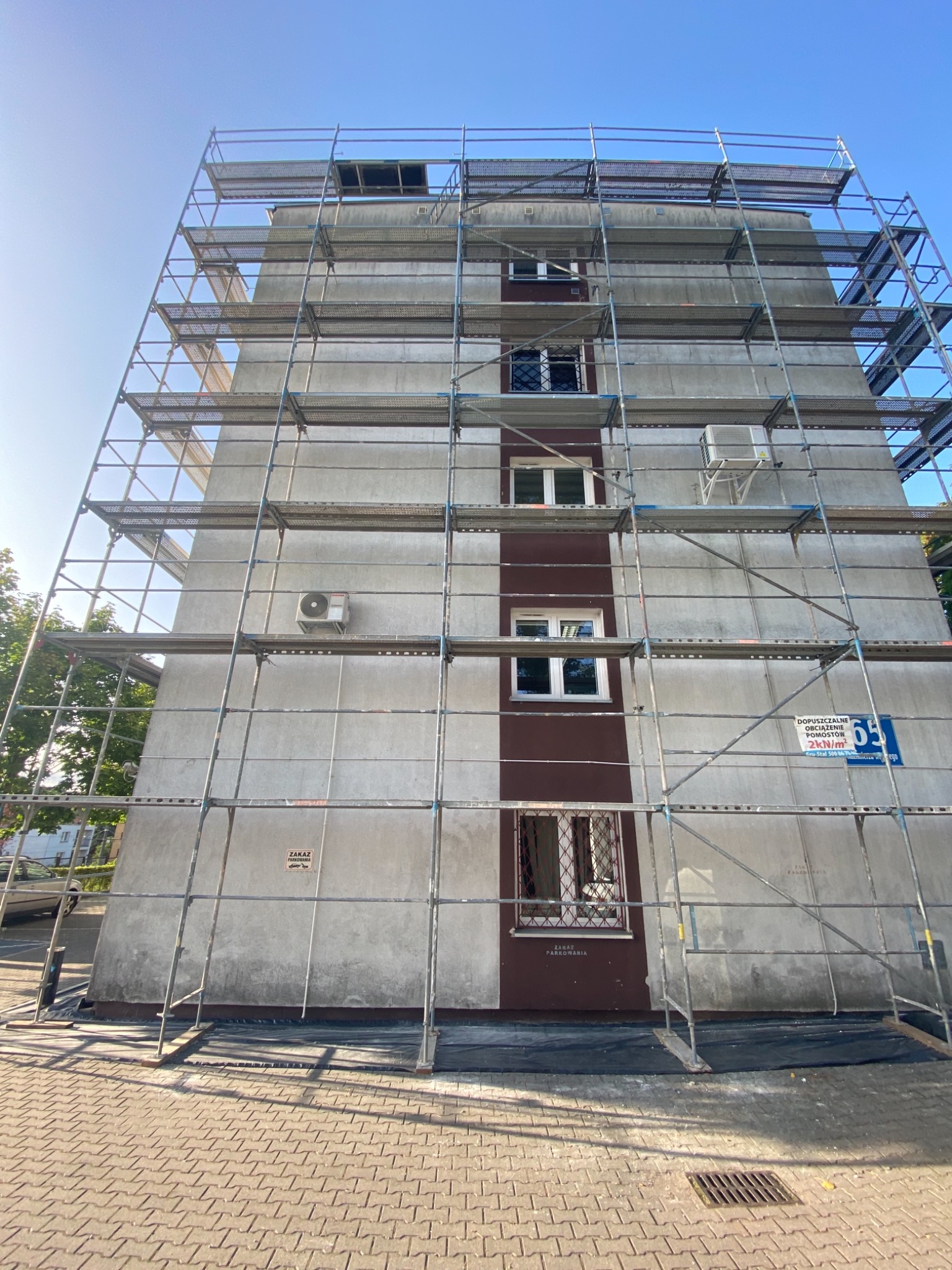 Wysoki budynek mieszkalny w trakcie prac elewacyjnych, otoczony rusztowaniem, widoczne okna, klimatyzator i tablica informacyjna z numerem 65.