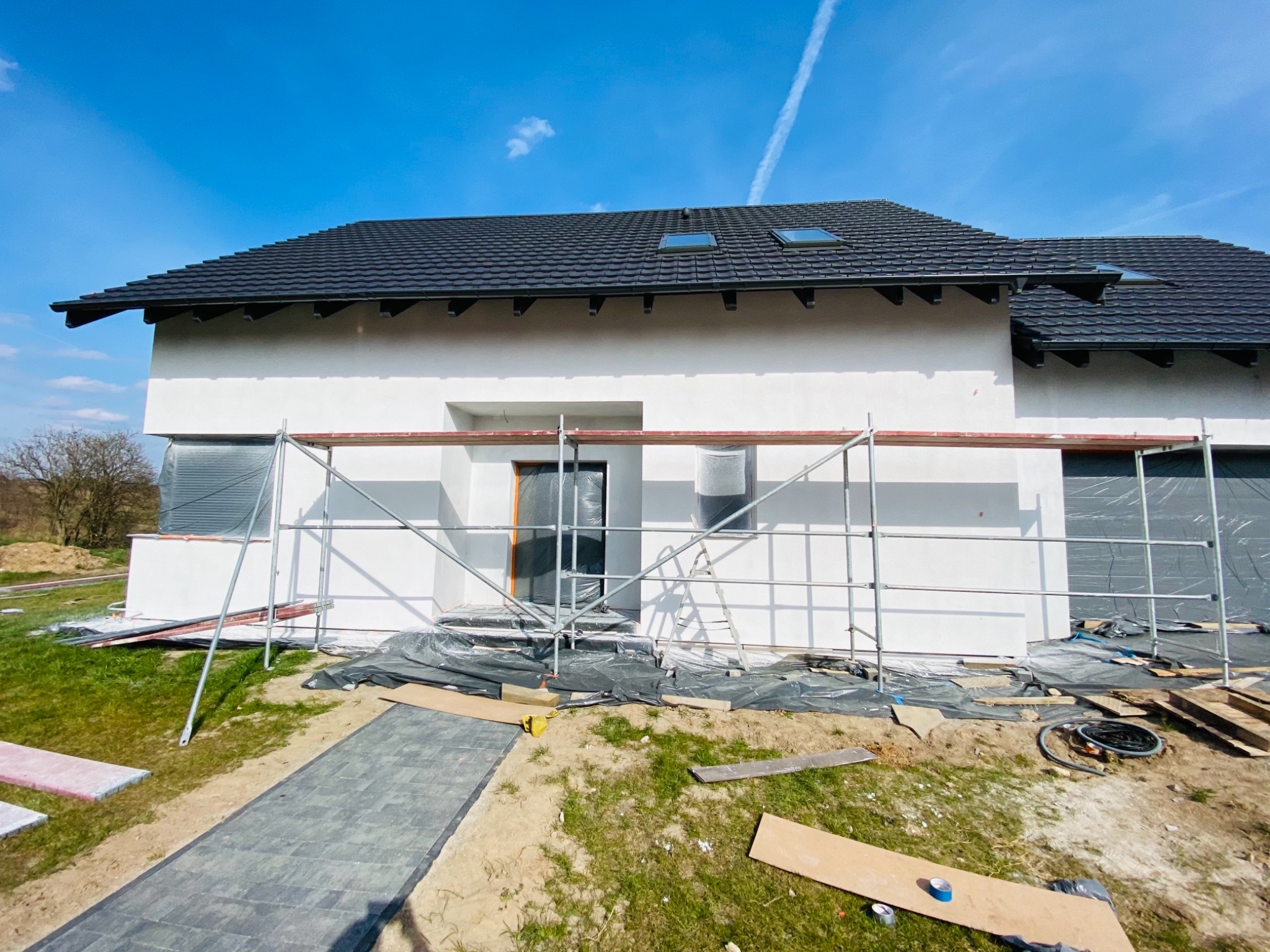 Biała elewacja domu jednorodzinnego w trakcie prac wykończeniowych, widoczne rusztowanie, folia ochronna na oknach i drzwiach, oraz częściowo ułożony chodnik z kostki brukowej. Na dachu czarna...