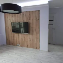 Telewizor zawieszony na ścianie wyłożonej panelami drewnopodobnymi w odcieniach brązu, podświetlony od góry listwą LED, z szarą podłogą imitującą drewno i czarną lampą wiszącą w tle.