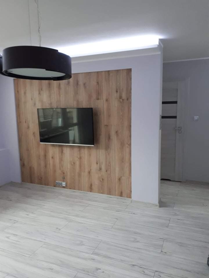 Telewizor zawieszony na ścianie wyłożonej panelami drewnopodobnymi w odcieniach brązu, podświetlony od góry listwą LED, z szarą podłogą imitującą drewno i czarną lampą wiszącą w tle.