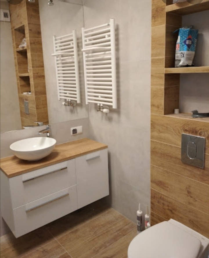 Nowoczesna łazienka z umywalką nablatową, szafką z drewnianym blatem, dwoma białymi grzejnikami drabinkowymi i płytkami imitującymi drewno na podłodze i ścianie.