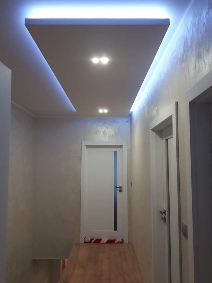 Korytarz z dekoracyjną strukturą na ścianach, podwieszany sufit z oświetleniem LED i punktowymi lampami, drewniana podłoga, białe drzwi z przeszkleniem.