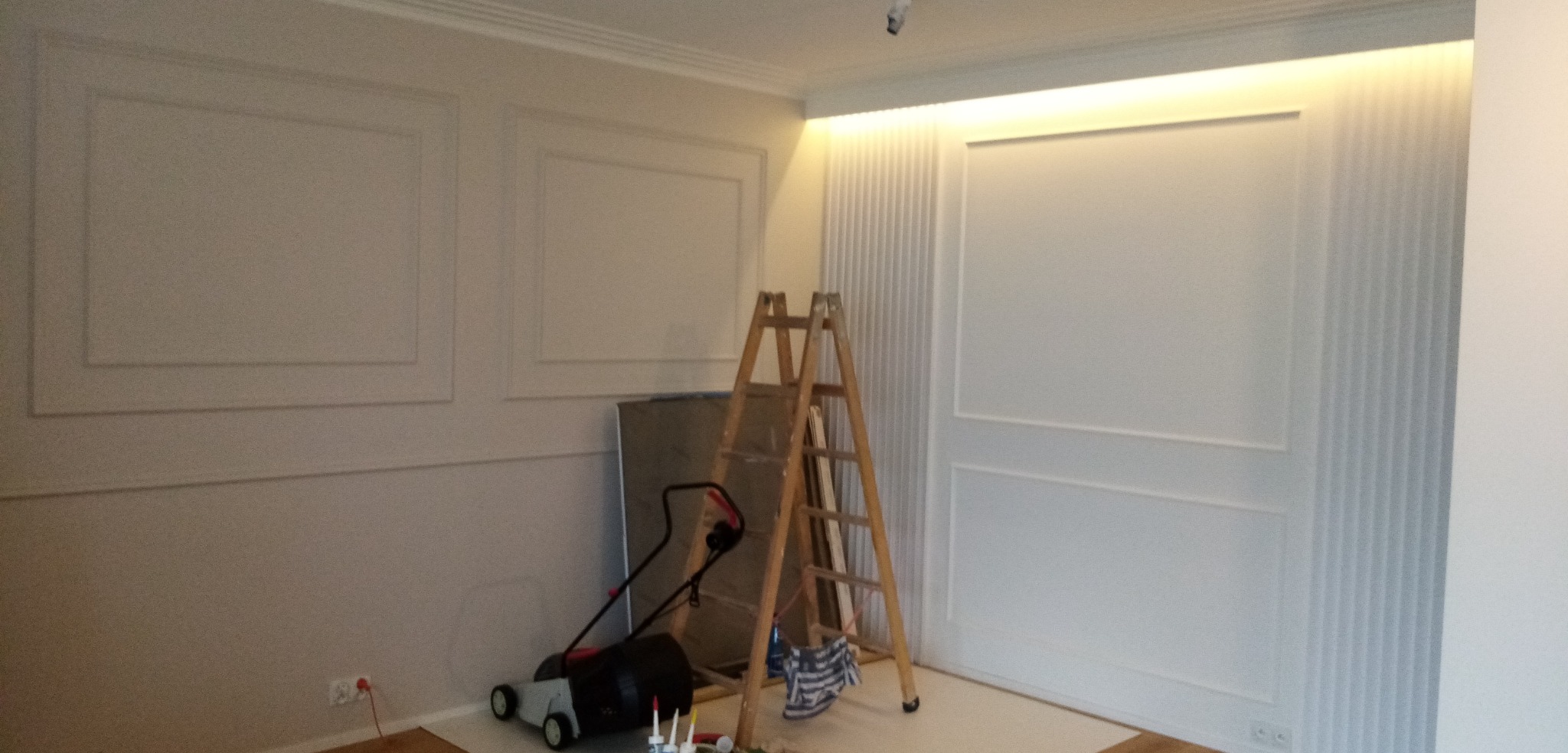 Świeżo pomalowane, białe ściany z dekoracyjnymi panelami, drabina oparta o ścianę i zakryta folią podłoga w trakcie prac wykończeniowych.