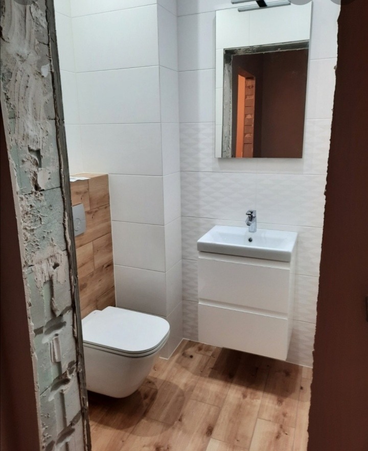 Nowoczesna łazienka z białymi płytkami, wiszącą toaletą i szafką z umywalką, drewnopodobna podłoga, widoczny fragment surowej ściany w trakcie remontu