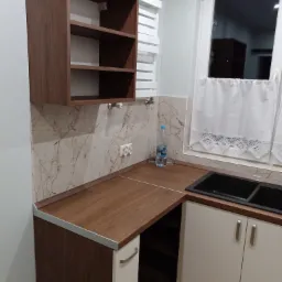 Nowoczesna zabudowa kuchenna w Łowiczu z drewnianym blatem, szafką wiszącą z otwartymi półkami i marmurowym panelem ściennym, uzupełniona o białe szafki dolne i czarny zlew.