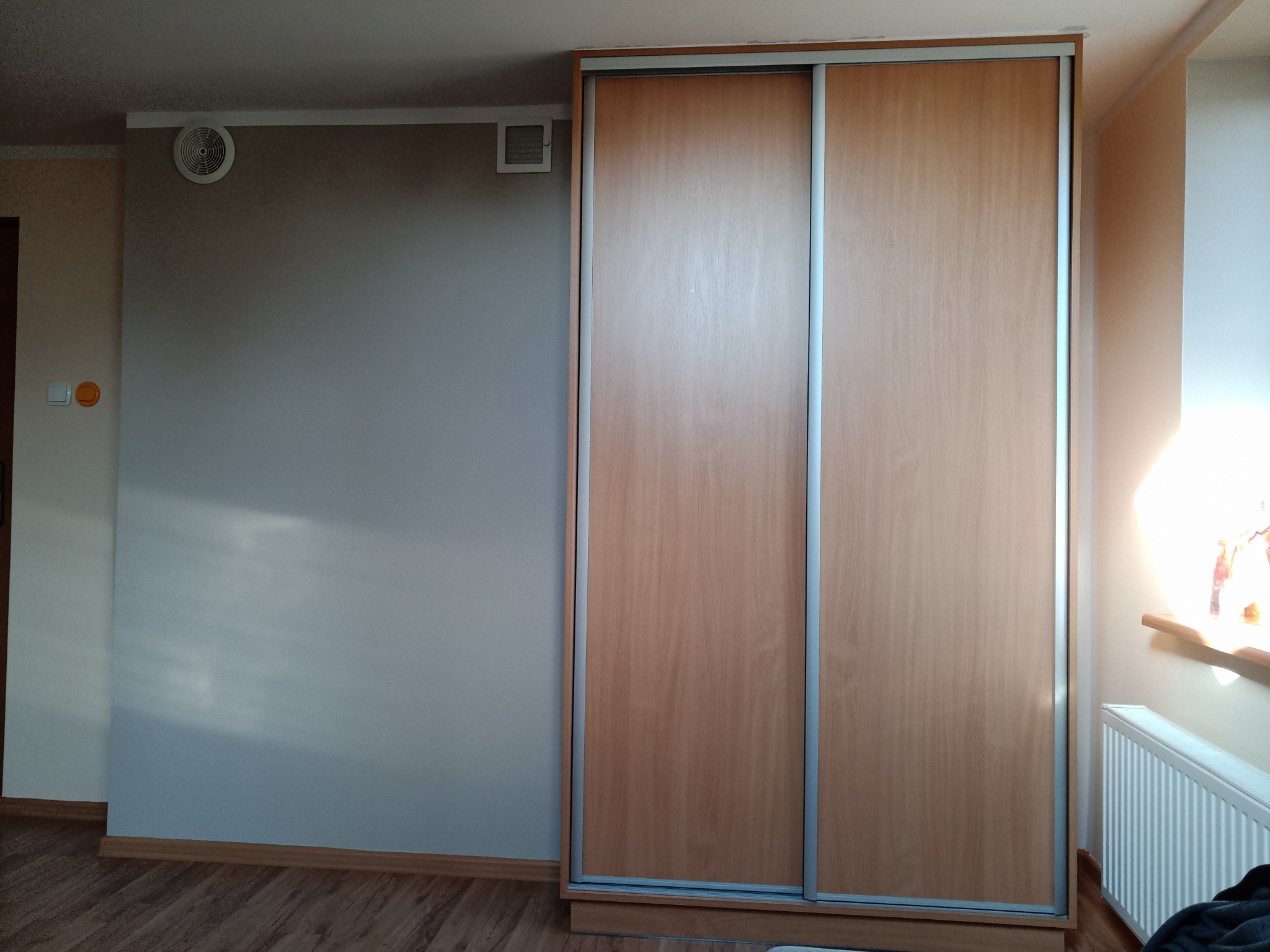 Beżowa szafa przesuwna z jasnymi, pionowymi, metalowymi uchwytami na tle szarej ściany z kratką wentylacyjną i okrągłym nawiewnikiem.