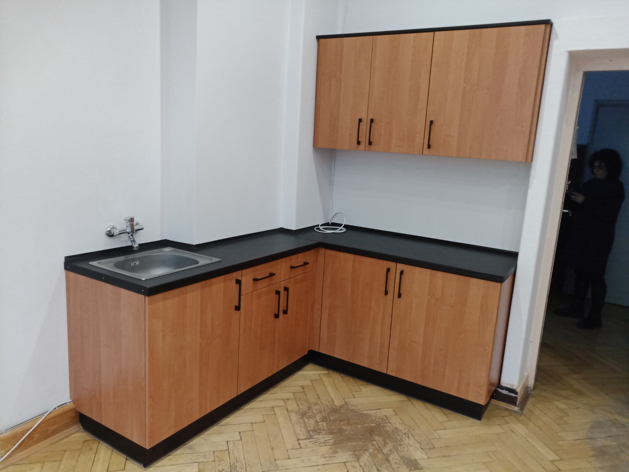 Kącik kuchenny z drewnianymi szafkami z czarnym blatem i zlewozmywakiem ze stali nierdzewnej, widoczne podłogi z parkietu i fragment sylwetki osoby w tle.