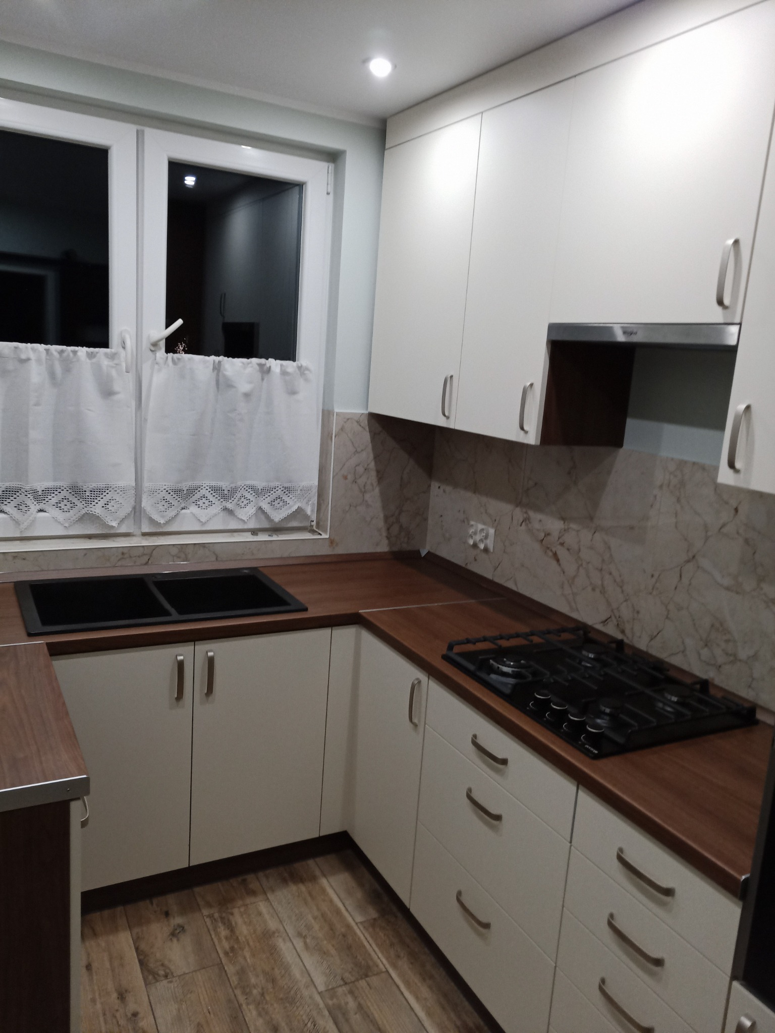 Nowoczesna kuchnia z białymi szafkami i blatem w kolorze drewna, czarny zlew pod oknem z koronkowymi firankami, płyta gazowa na blacie, brązowe płytki imitujące marmur na ścianie.