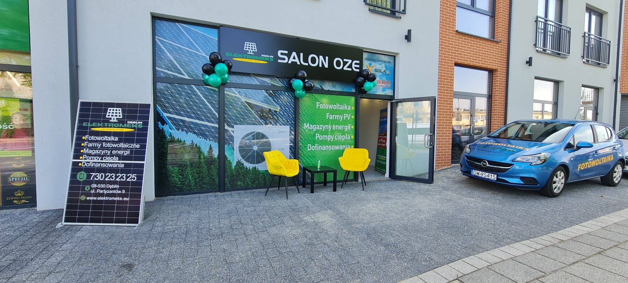 Salon OZE firmy Elektromeks w Dęblinie, udekorowany balonami, z widoczną ofertą fotowoltaiki, pomp ciepła i dofinansowań. Na parkingu stoi firmowy Opel z napisem Fotowoltaika.