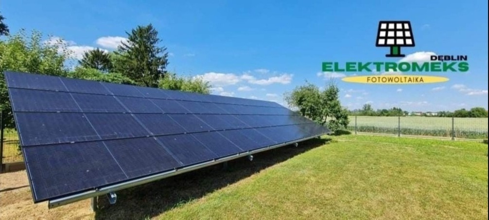 Nisko osadzona instalacja paneli słonecznych na trawniku, widoczna marka ELEKTROMEKS Dęblin, w tle zielone drzewa i niebieskie niebo.