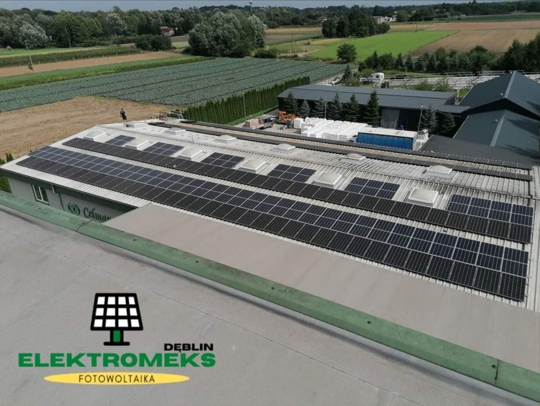 Widok z góry na dach budynku z zamontowanymi panelami słonecznymi, logo firmy instalacyjnej w rogu, pola uprawne w tle.