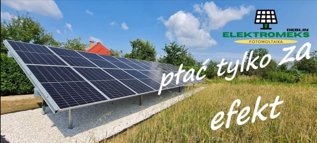 Instalacja paneli słonecznych na konstrukcji wsporczej, widoczna na tle wysokiej trawy i błękitnego nieba z chmurami. W tle budynek z czerwonym dachem i logo firmy Elektromeks Dęblin.