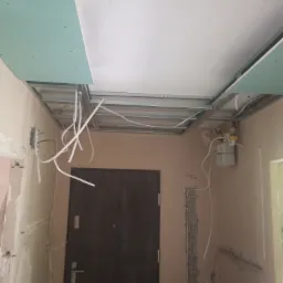 Montaż stelaża sufitu podwieszanego z płyt kartonowo-gipsowych, widoczne kable elektryczne i instalacja gazowa, ściany w trakcie przygotowania.