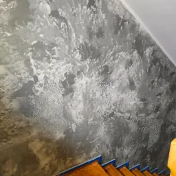 Ściana w szaro-srebrnej tonacji z dekoracyjną strukturą i subtelnym wzorem mandali, widoczne drewniane schody z niebieskimi akcentami.