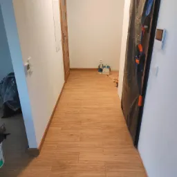 Korytarz po remoncie z nową, drewnopodobną podłogą z płytek. Zabezpieczone drzwi folią malarską, widoczne narzędzia i wiaderka w tle. Jasne ściany, drewniane listwy przypodłogowe.