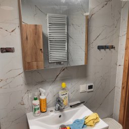 Tobiasz Madoń - Wykończona łazienka z marmurowymi płytkami. Widoczne lustro, szafka z drewnianymi frontami, grzejnik drabinkowy i umywalka z akcesoriami.