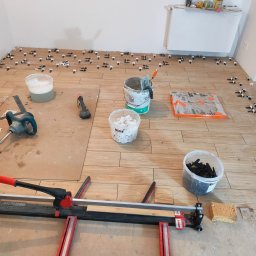 Tobiasz Madoń - Układanie płytek podłogowych z użyciem klipsów poziomujących i przecinarki RedTechnic. Widoczne narzędzia glazurnicze i materiały budowlane w jasnym pomieszczeniu.