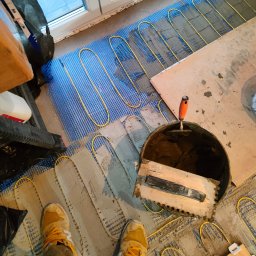 Tobiasz Madoń - Montaż ogrzewania podłogowego: żółte kable grzewcze na niebieskiej siatce, częściowo pokryte zaprawą, wiadro z narzędziami, widok z góry, buty pracownika.