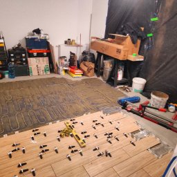 Tobiasz Madoń - Układanie płytek podłogowych na ogrzewaniu podłogowym. Widoczny poziomica i narzędzia glazurnicze. Częściowo ułożone płytki z krzyżykami dystansowymi.