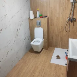 Wykończona łazienka z drewnopodobnymi i marmurowymi płytkami, wisząca toaleta, częściowo widoczna umywalka i armatura prysznicowa. W tle akcesoria glazurnicze.