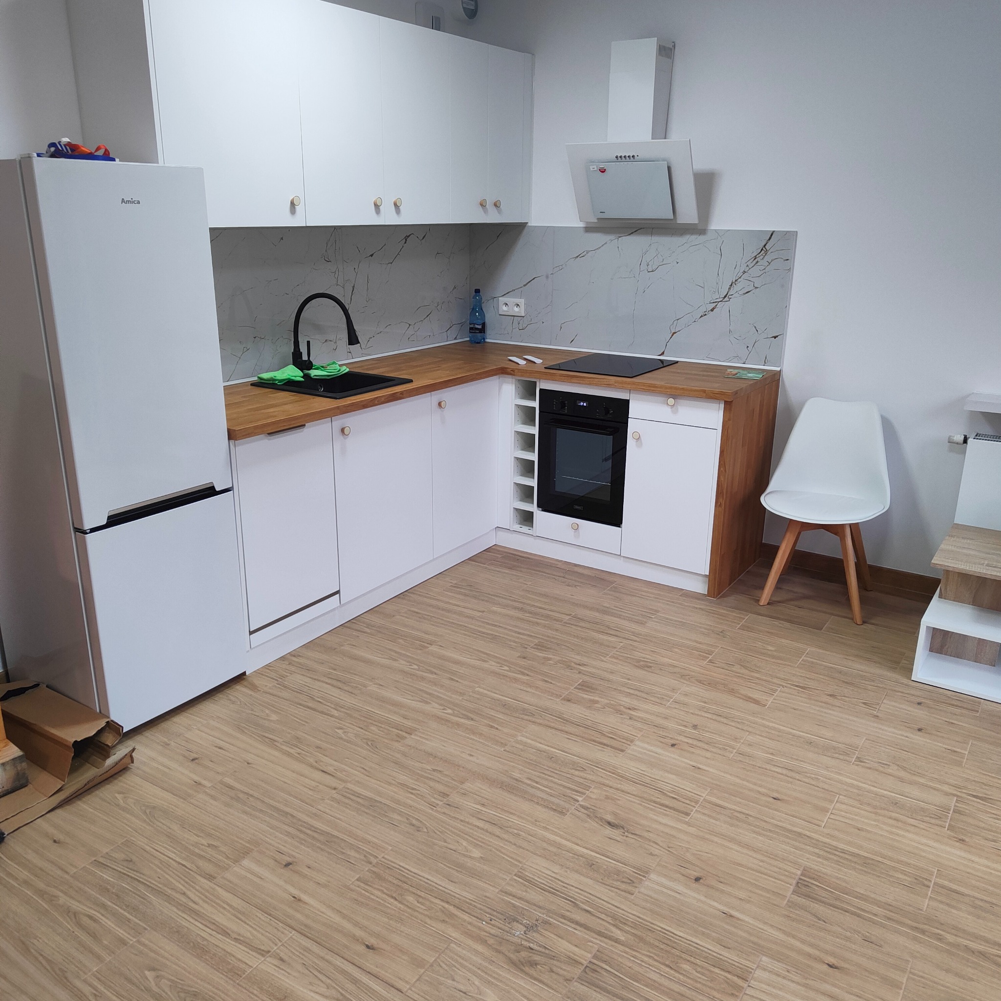 Jasna kuchnia z białymi szafkami i drewnianym blatem. Na podłodze płytki imitujące drewno. Nowoczesny design z czarną armaturą i płytą indukcyjną.