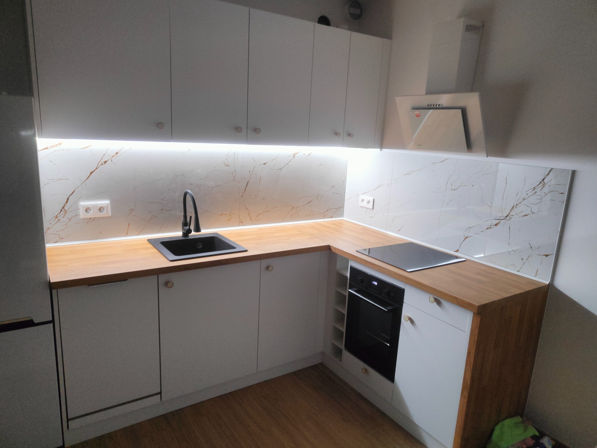 Nowoczesna kuchnia w bieli i drewnie: zabudowa z oświetleniem LED, marmurowe panele ścienne i czarny zlew. Minimalistyczny design z elementami naturalnego drewna.