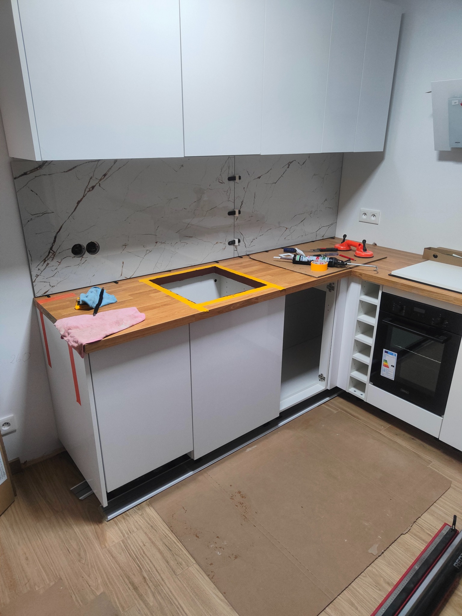 Montaż blatu i płytek imitujących marmur w nowoczesnej kuchni. Widoczne narzędzia i akcesoria montażowe, płyta indukcyjna jeszcze nie zamontowana.