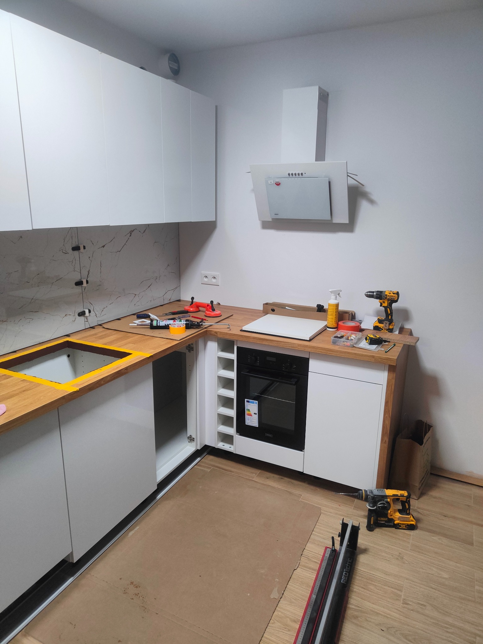 Montaż kuchni: białe szafki, drewniany blat, sprzęt AGD. Widoczne narzędzia i materiały, fragment marmurowej ściany. Ujęcie z bliska na etapie prac.