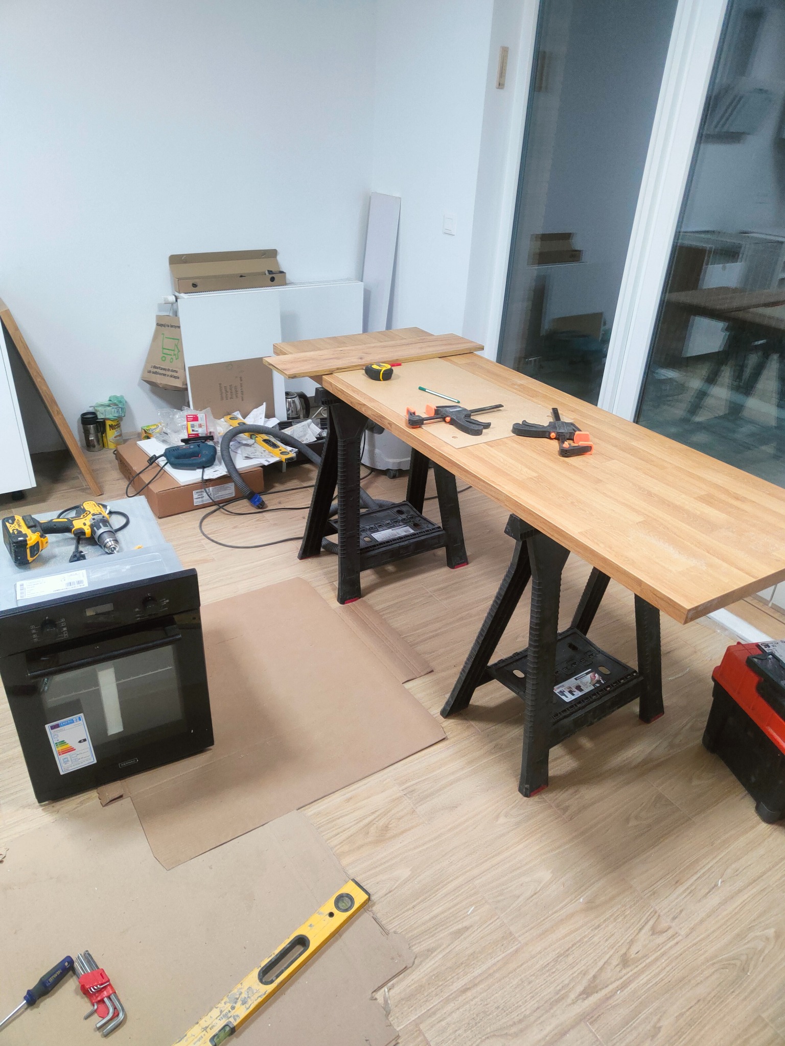 Montaż blatu kuchennego: drewniany blat na kozłach, narzędzia, poziomica, w tle piekarnik i okno balkonowe. Etap prac wykończeniowych w jasnym wnętrzu.