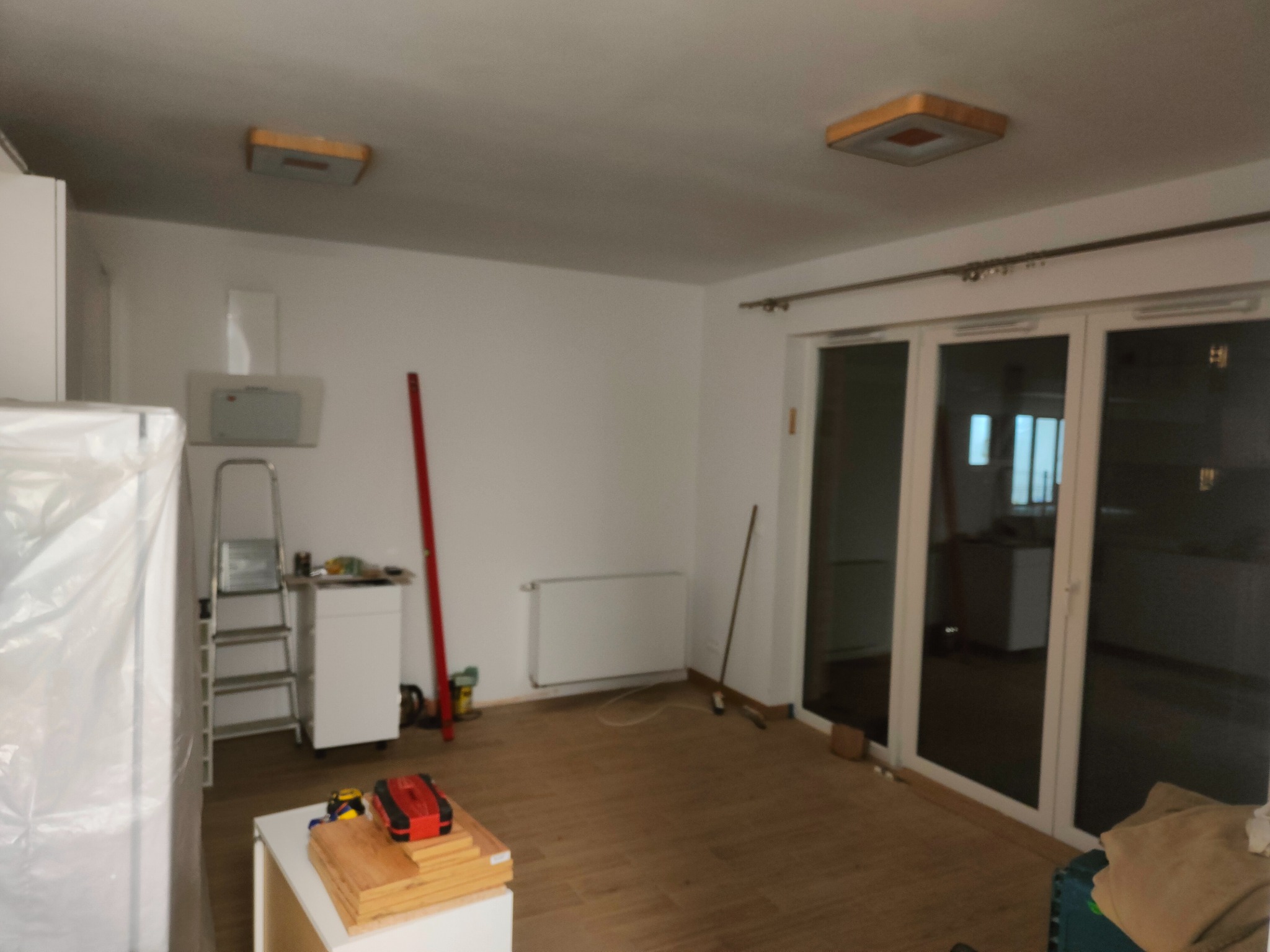 Jasne wnętrze w trakcie remontu: parkiet, białe ściany, drabina, narzędzia i okno balkonowe. Widoczne przygotowania do prac wykończeniowych.