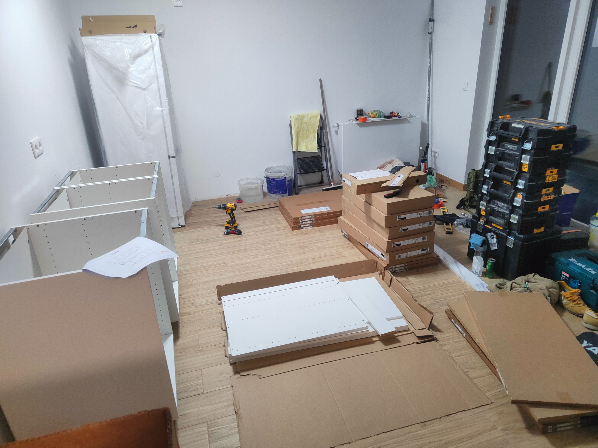 Wnętrze w trakcie montażu mebli kuchennych: rozpakowane elementy, skrzynki narzędziowe, kartony i instrukcja na podłodze, sprzęt AGD w folii przy ścianie.