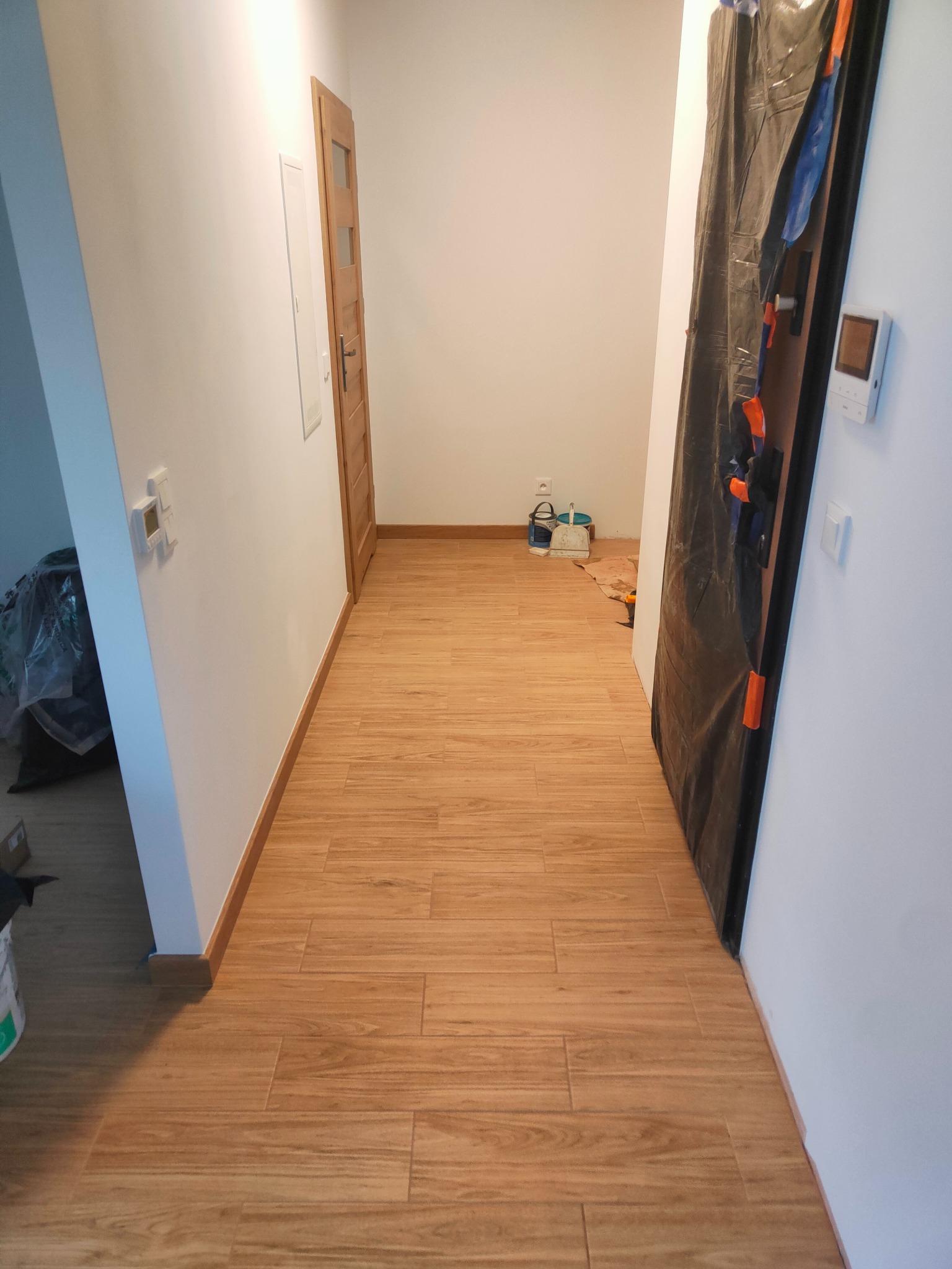 Korytarz po remoncie z nową, drewnopodobną podłogą z płytek. Zabezpieczone drzwi folią malarską, widoczne narzędzia i wiaderka w tle. Jasne ściany, drewniane listwy przypodłogowe.